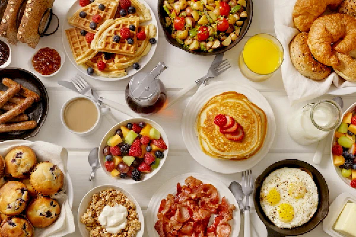 An assorted breakfast spread: waffles, pancakes, fruits, eggs, bacon, croissants, muffins, granola, juice, coffee, and syrup.