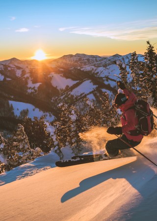 A skier is descending a snowy slope at sunset, surrounded by pine trees and a mountainous landscape.