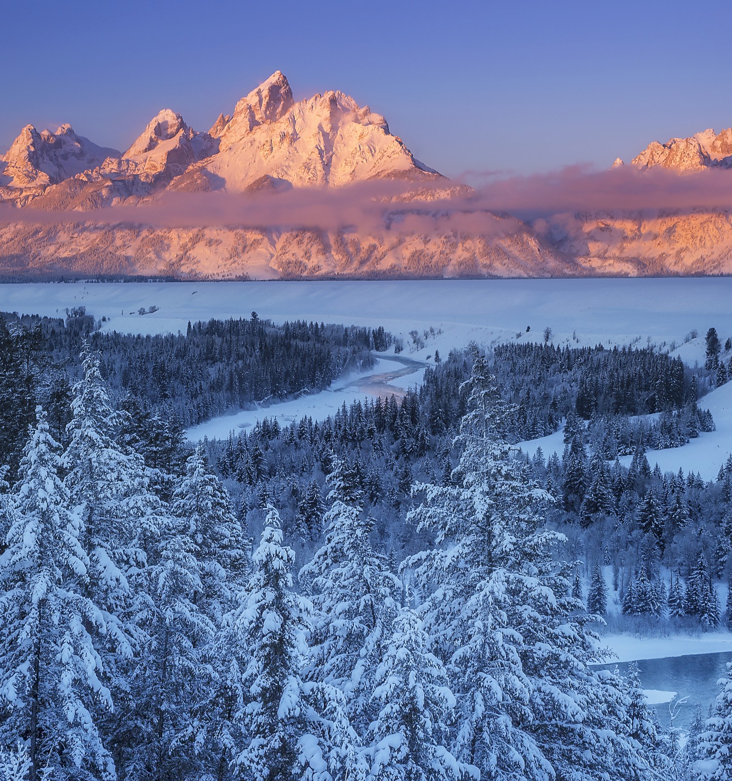 A winter landscape with snow-covered trees and mountains at sunset, casting an orange glow. A river winds through the snowy scene.