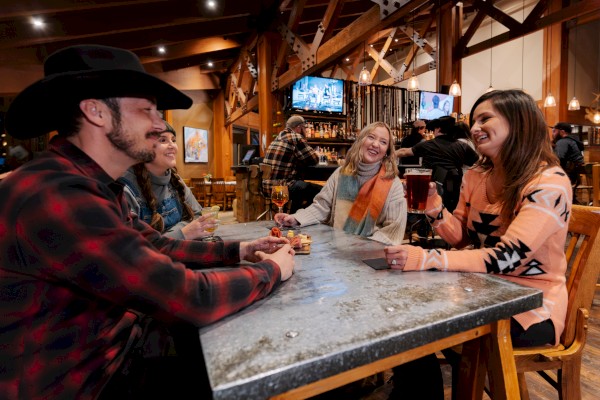 A group of people sitting at a table in a rustic setting, talking and enjoying drinks, with a TV and bar in the background.