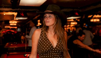 A person in a polka dot dress and hat stands next to a pool table in a dimly lit room with warm lights and people in the background.