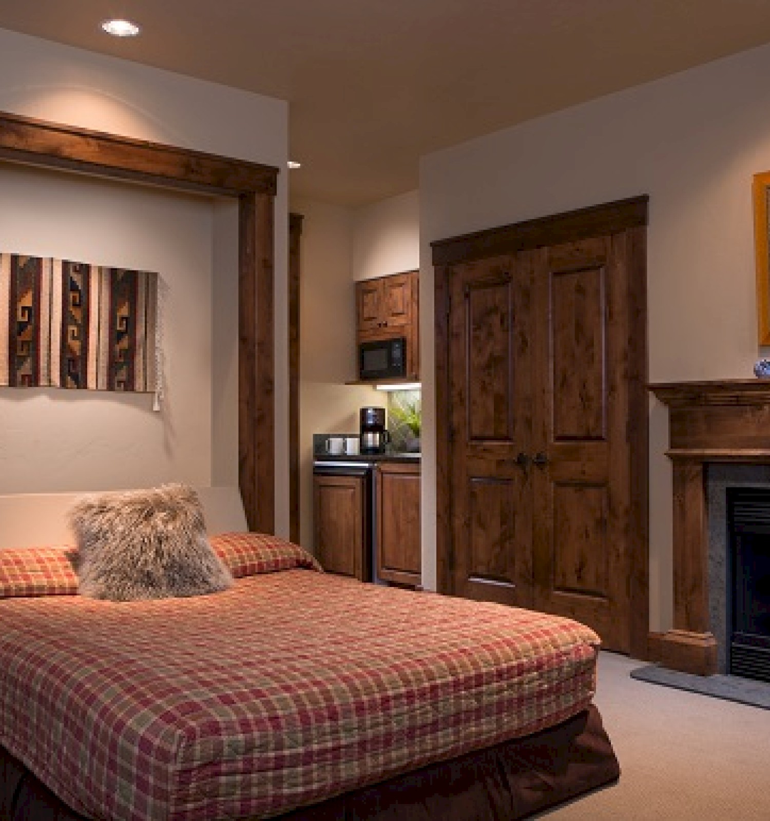 The image shows a cozy bedroom with a plaid bed, a furry pillow, wooden accents, a small kitchen area, and a fireplace.