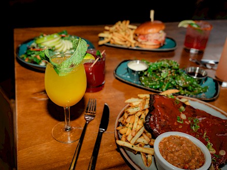 A table with cocktails, a salad, a burger with fries, ribs with fries and beans, and greens, all artfully arranged for dining.