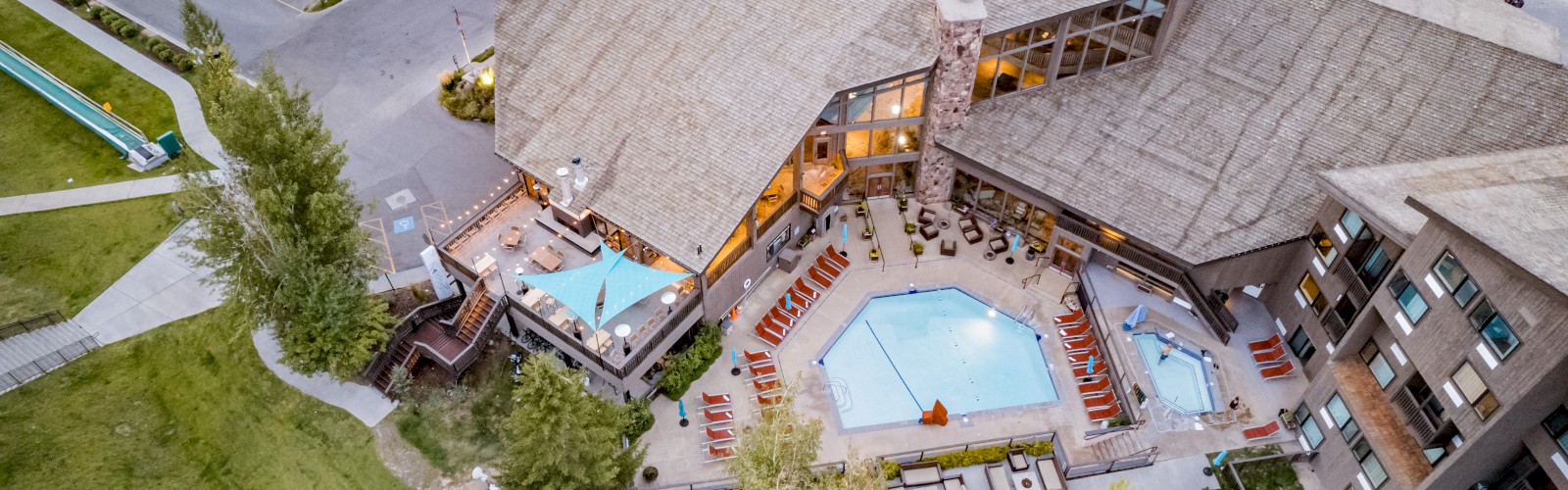 An aerial view of a building with two outdoor swimming pools surrounded by lounge chairs, adjacent to a parking lot and landscaped greenery.