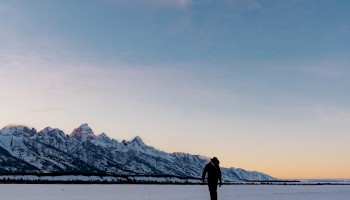 A person stands on a vast snowy field with distant mountains and a colorful sky at sunset or sunrise.