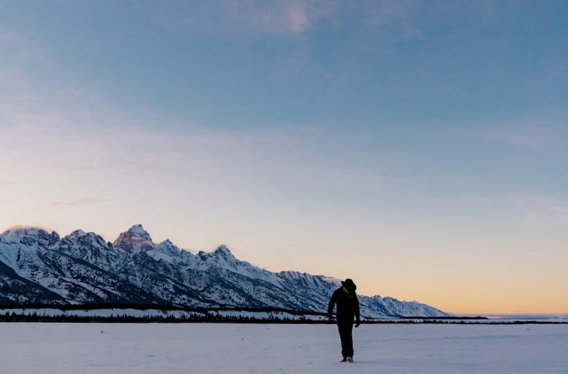 A person stands on a vast snowy field with distant mountains and a colorful sky at sunset or sunrise.
