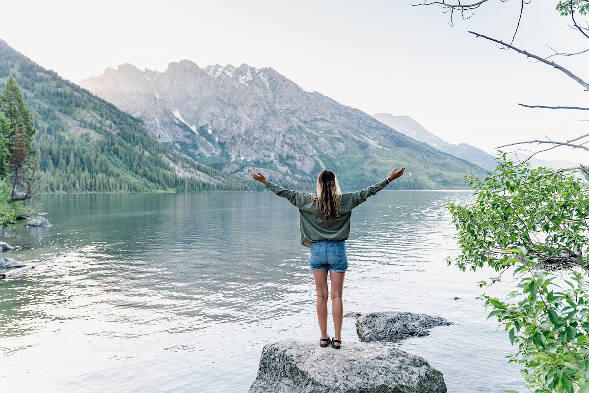 A person stands on a rock by a lake with arms raised, surrounded by mountains and greenery.