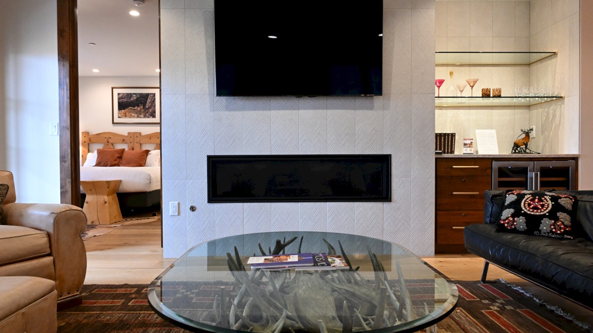 A cozy living room with a TV, fireplace, and glass table. Bedroom and shelf with decor are visible in the background.