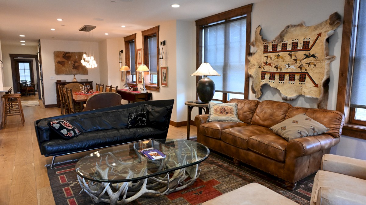The image shows a cozy living room with leather sofas, a glass table, and decorative wall art, adjacent to a dining area.