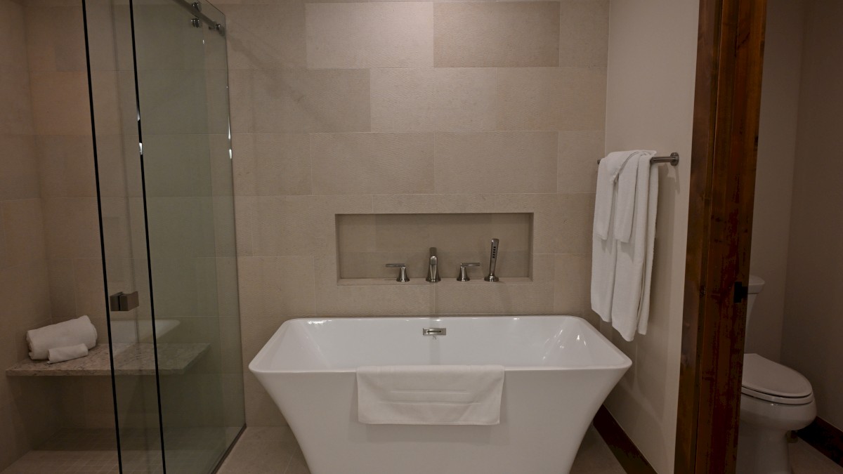 A modern bathroom with a glass shower, freestanding bathtub, and toilet, all set against beige tiles, with towels on racks.