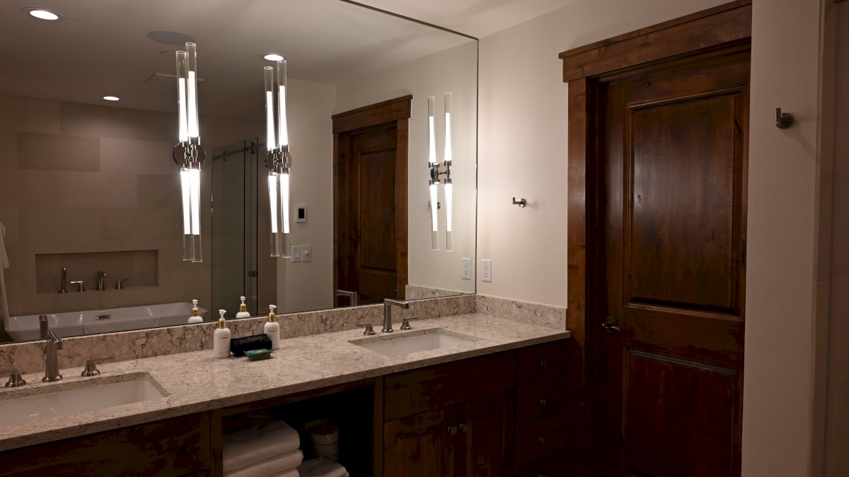 A modern bathroom with a double sink vanity, large mirror, elegant lighting, wooden door, and towels neatly stacked below.