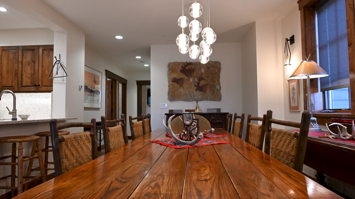 A dining room with a long wooden table, chairs, a central light fixture, wall decor, and a view into the kitchen area, completes the scene.