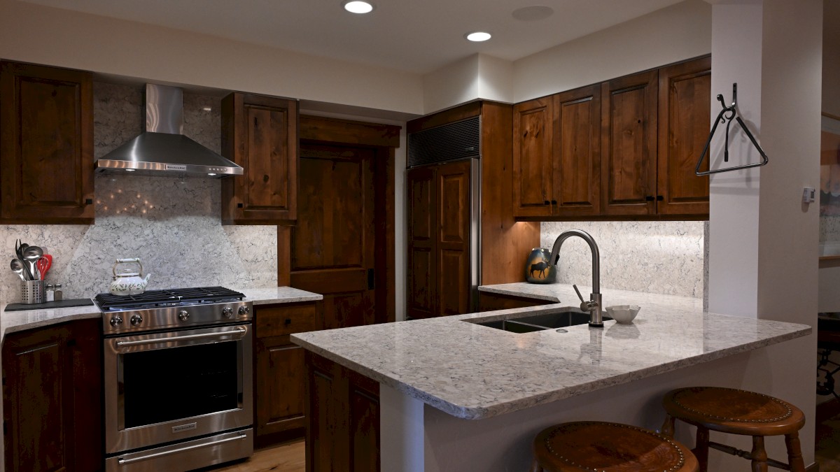 A modern kitchen with wooden cabinets, a stainless steel stove, marble countertops, and two stools at a breakfast bar.