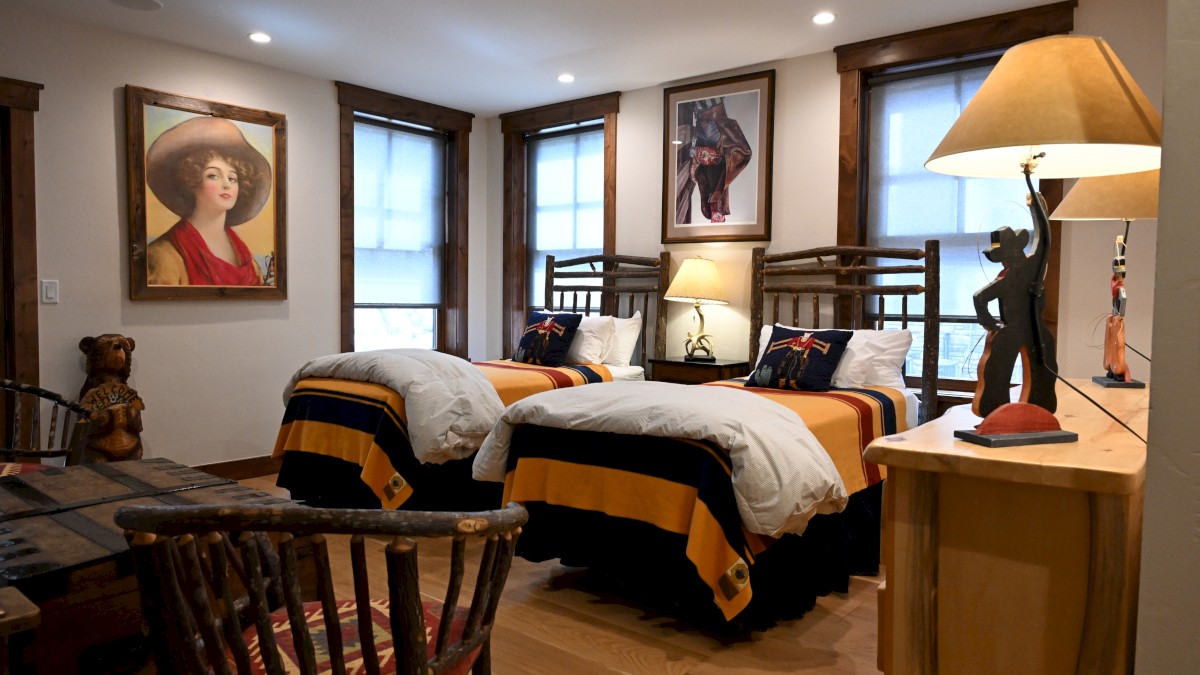 A cozy bedroom features two twin beds with striped blankets, artwork on the walls, a wooden chair, and soft lighting from a lamp.