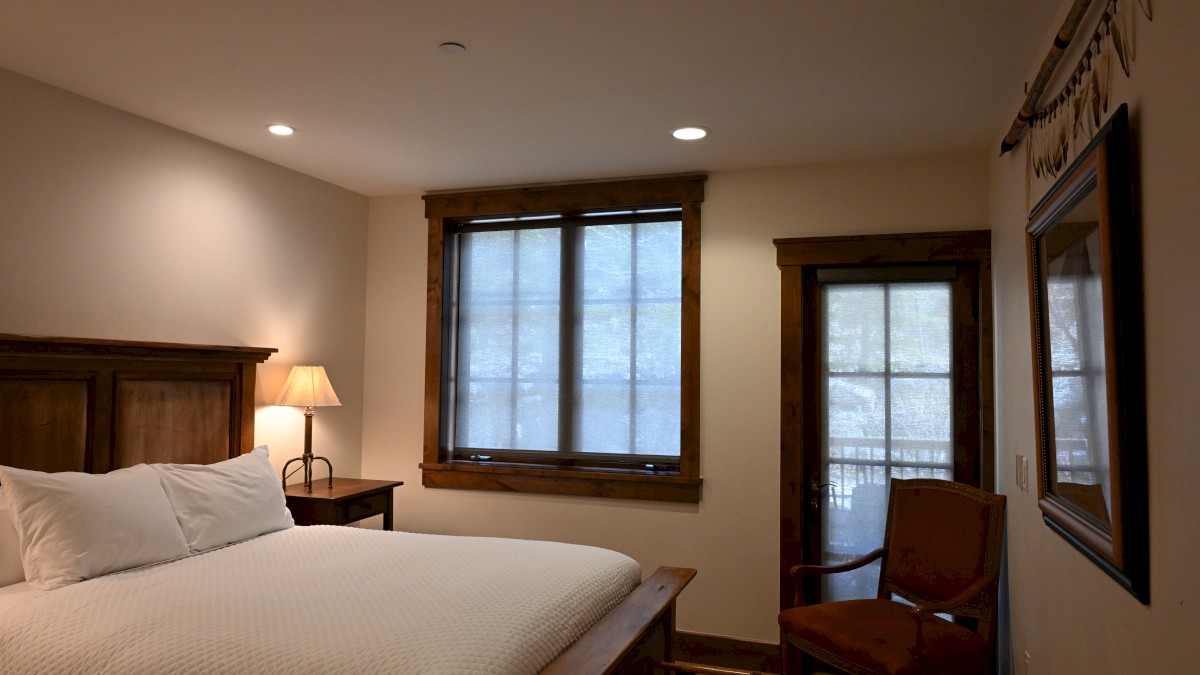 A cozy bedroom with a large bed, bedside lamp, window with blinds, wooden chair, and mirror on the wall, creating a warm atmosphere.