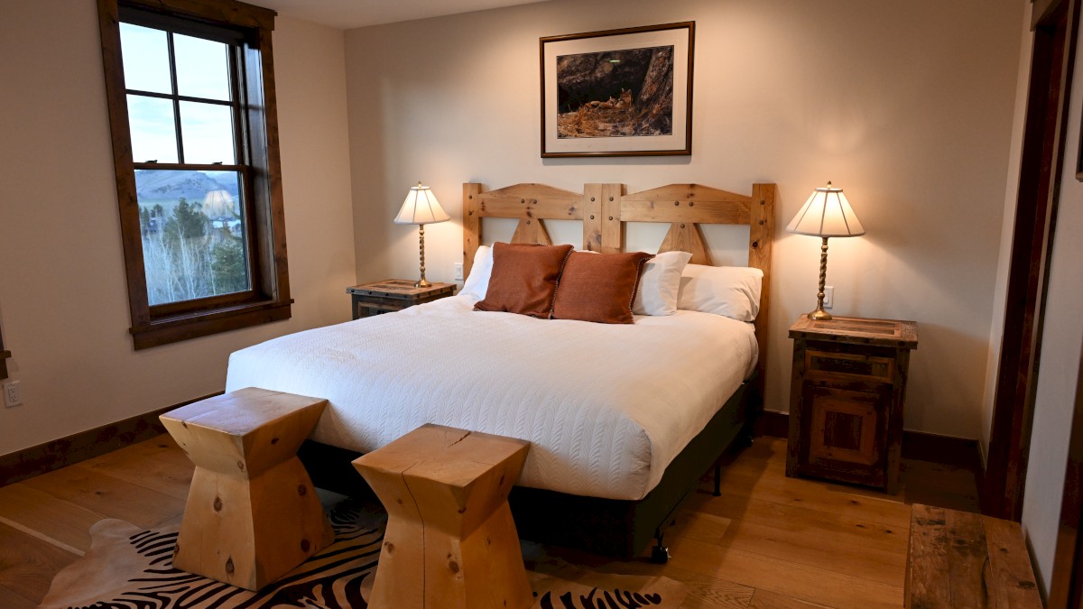 A cozy bedroom features a large bed, wooden furniture, bedside lamps, a window with a view, and a zebra-patterned rug on a wooden floor.