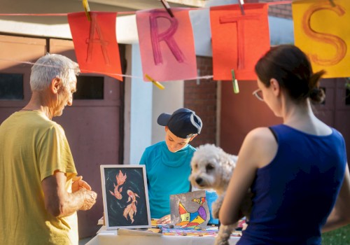 A small art stall is set up outdoors with paintings displayed. People are browsing, and a woman holds a dog. A banner reads 