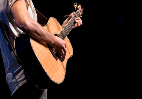 A person playing an acoustic guitar on stage, wearing a short-sleeved shirt, with a dark background, focusing on the instrument.