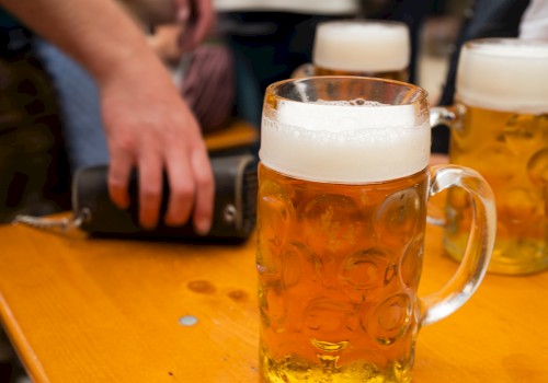 Mugs of beer with frothy heads are on a wooden table. A person is reaching for an object nearby.