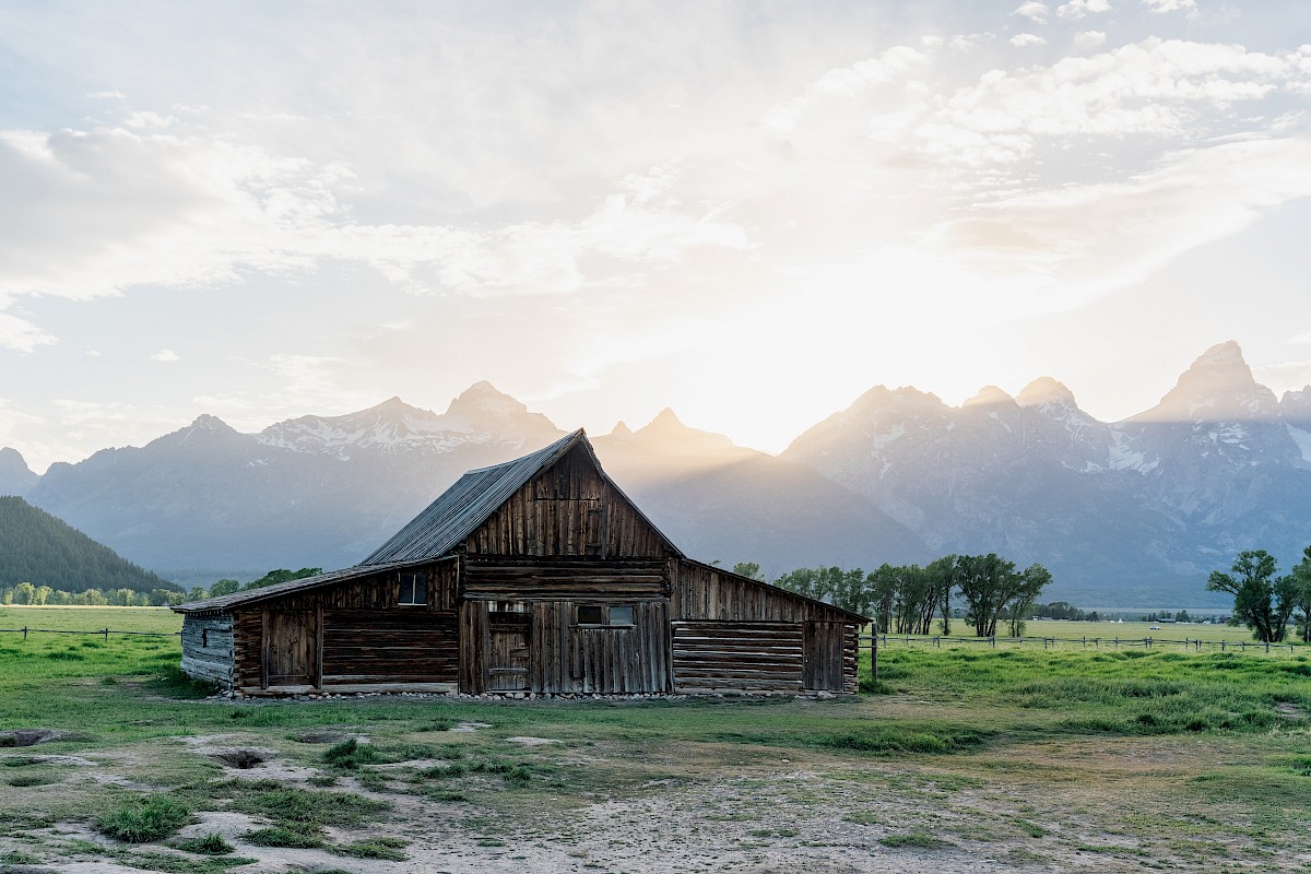A rustic wooden barn is set against a backdrop of mountains with the sun rising or setting, illuminating the landscape.