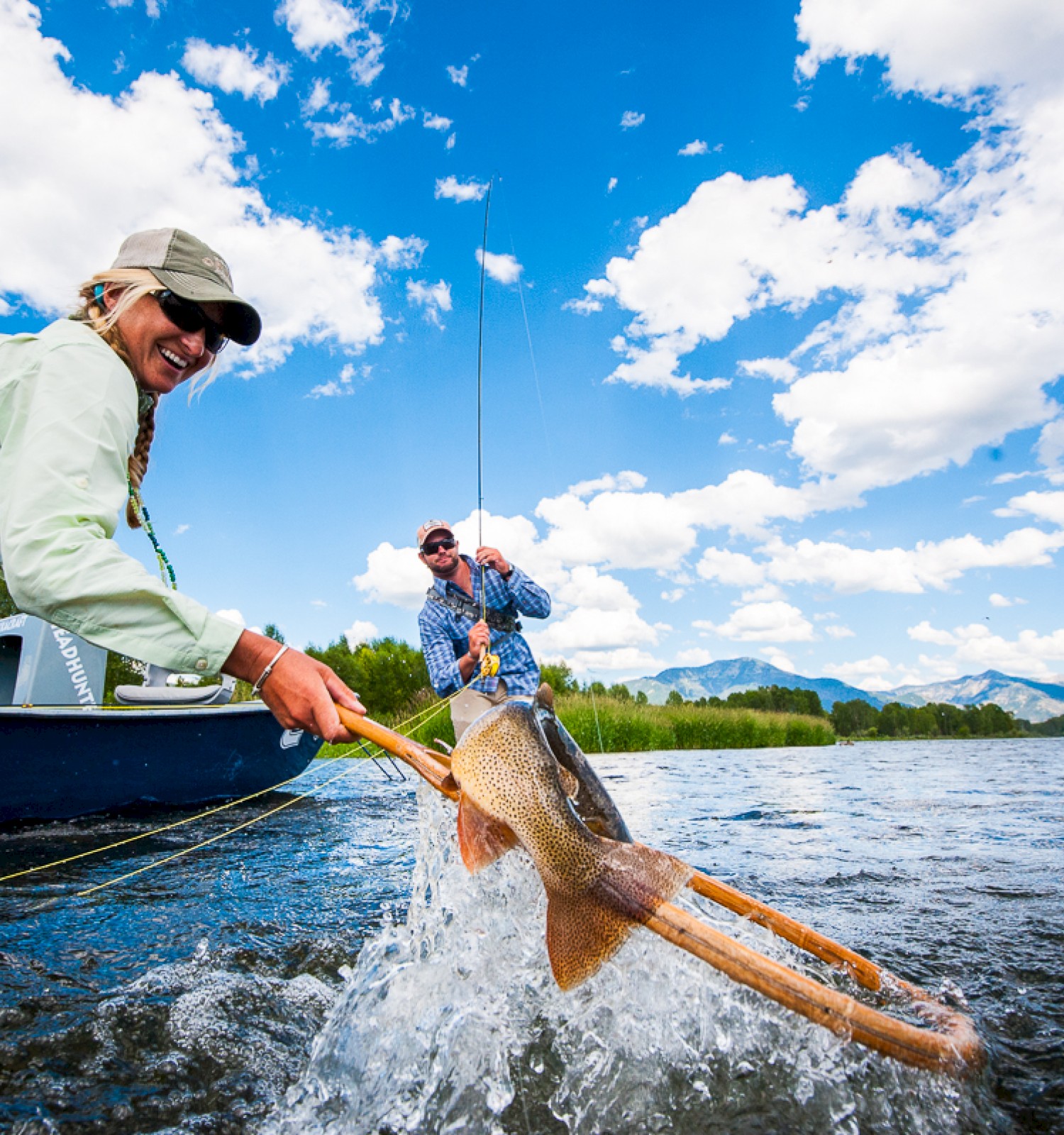 Two people fishing in a river; one holds a net catching a fish, while the other is in a boat, under a bright, partly cloudy sky.