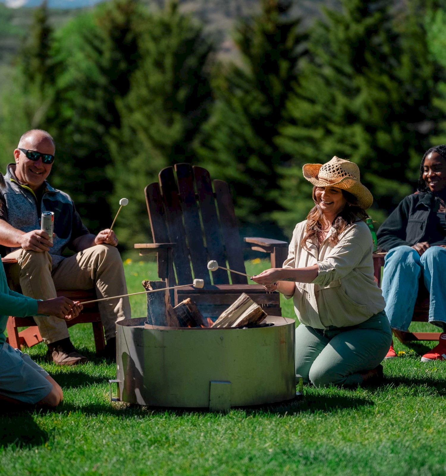 A group of people sit around a fire pit outdoors, roasting marshmallows and enjoying each other's company in a green, wooded area.