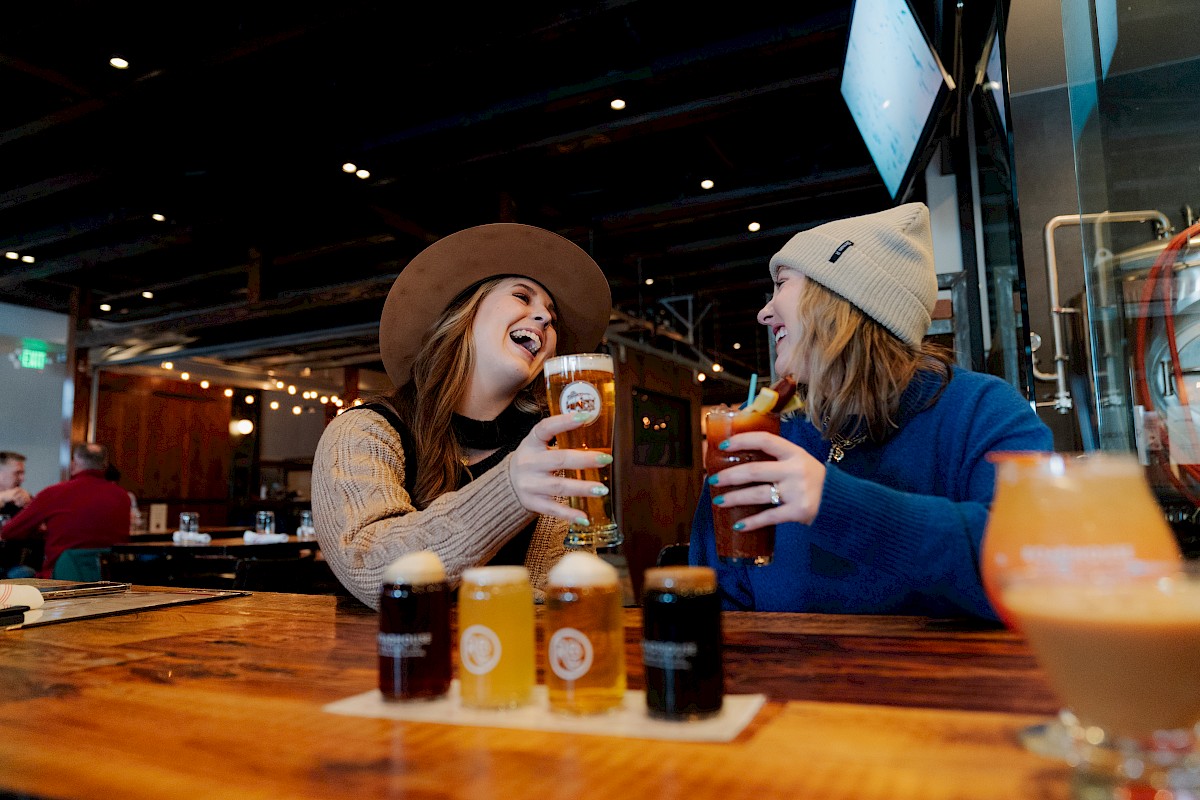 Two people enjoy drinks at a bar, laughing and holding beverages, with a flight of beers on the wooden table in front of them.