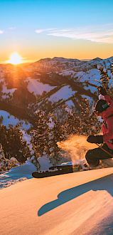 A skier glides down a snowy slope at sunset, powder spraying as trees line the ridge and orange light paints the mountains in the background.