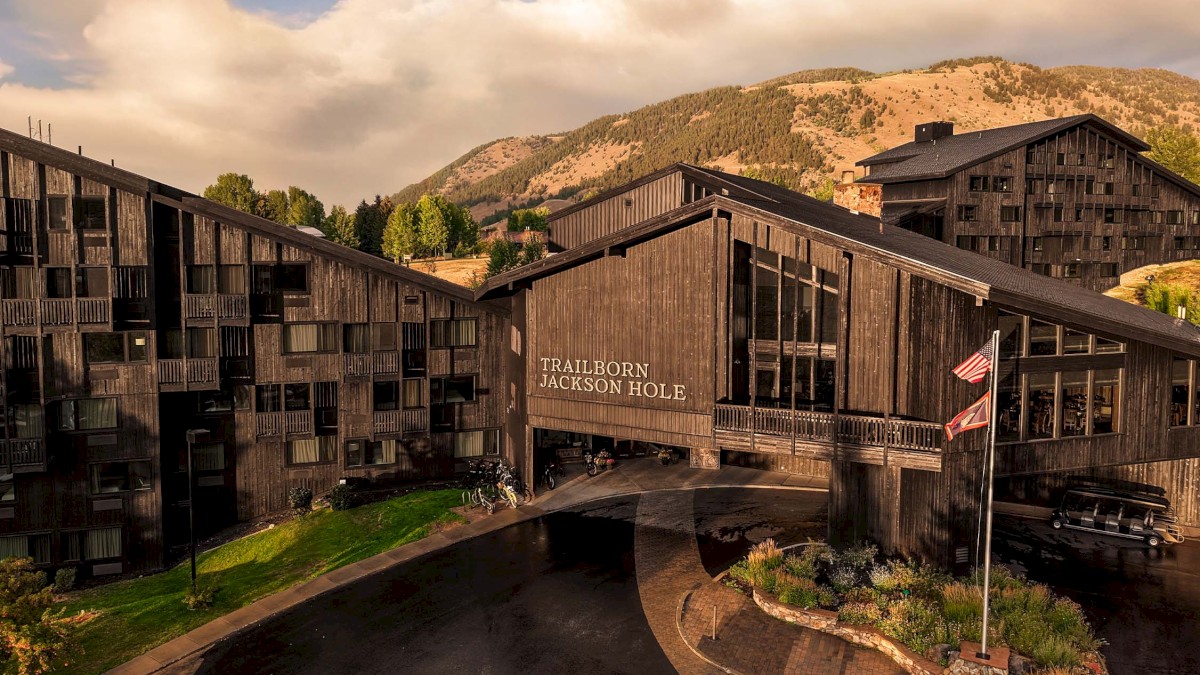 A rustic, mountain resort with dark wooden buildings connected by a central canopy bridge, set against rolling hills and a bright sky.