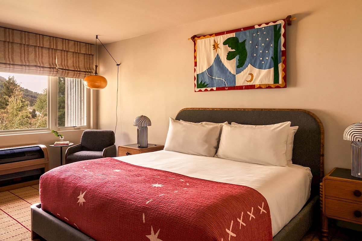A cozy hotel bedroom with a large bed, red throw blanket, two wooden nightstands, a window view, and a colorful wall tapestry above the headboard.