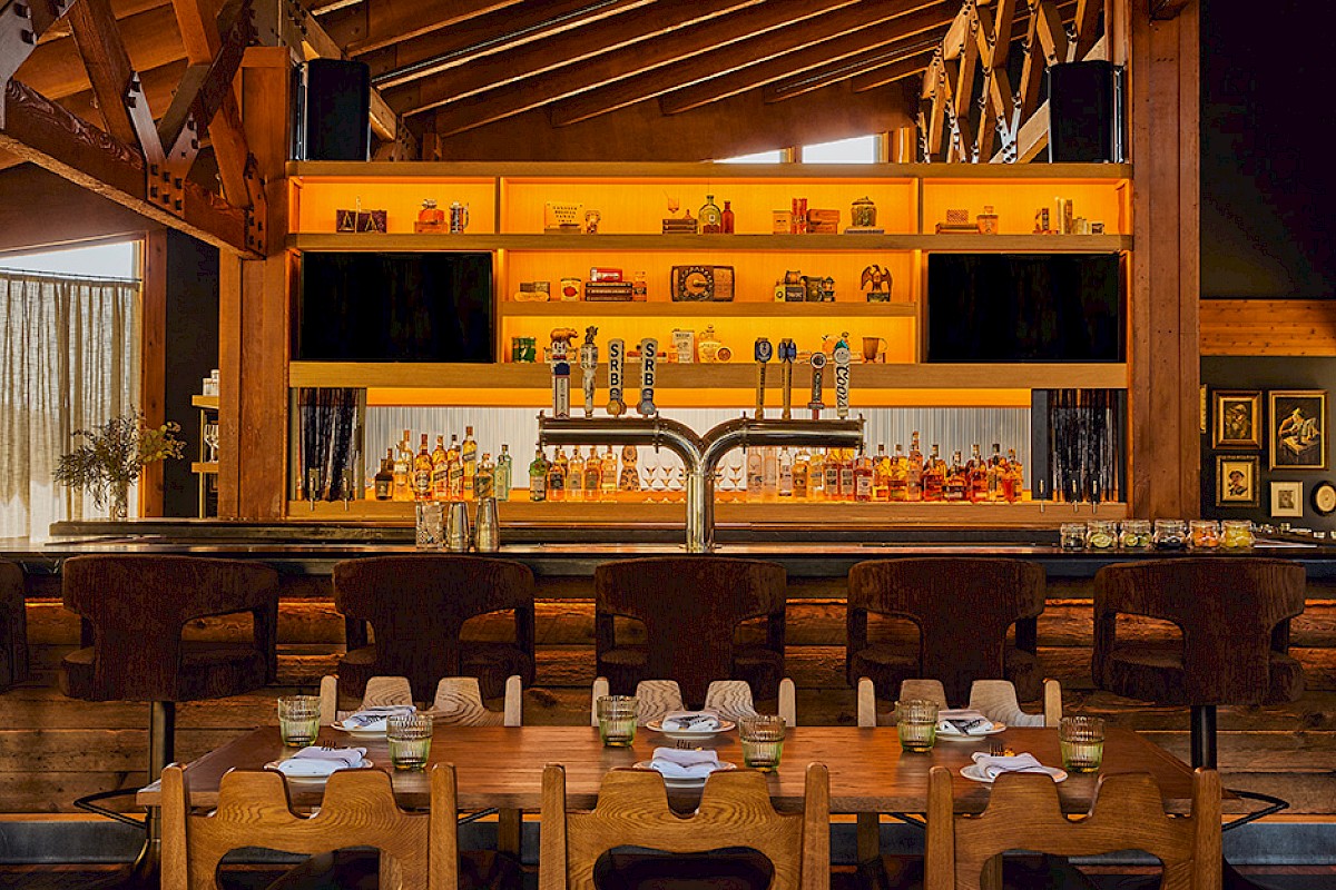 A cozy bar and dining area with a wooden interior, illuminated shelves with bottles, and a long table set for guests.