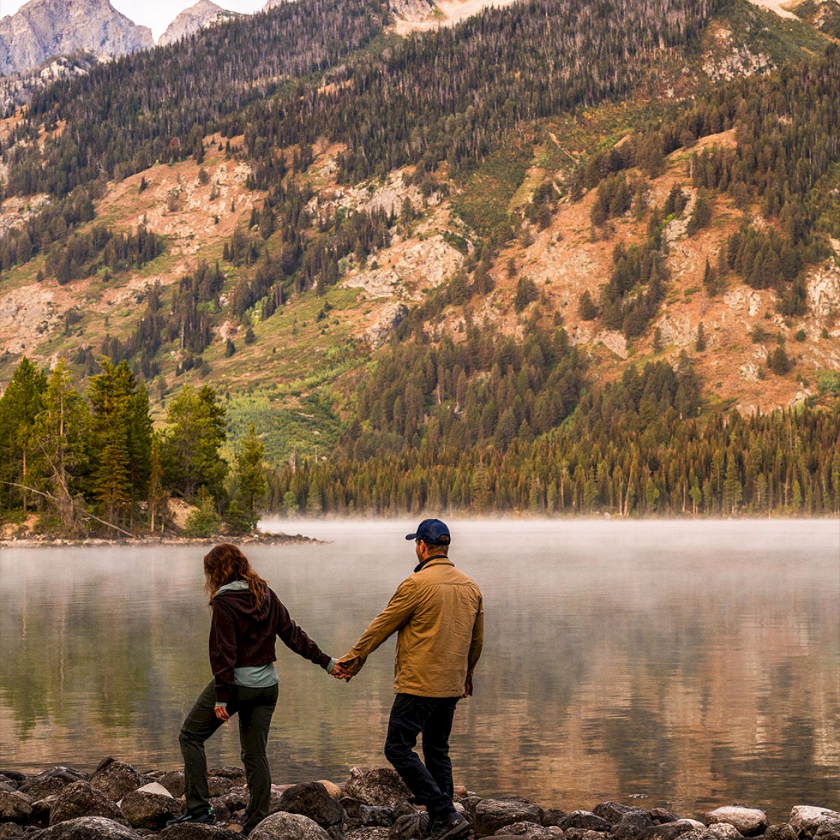 Two people holding hands by a misty lake, mountains and trees in the background, a peaceful outdoor couple moment.