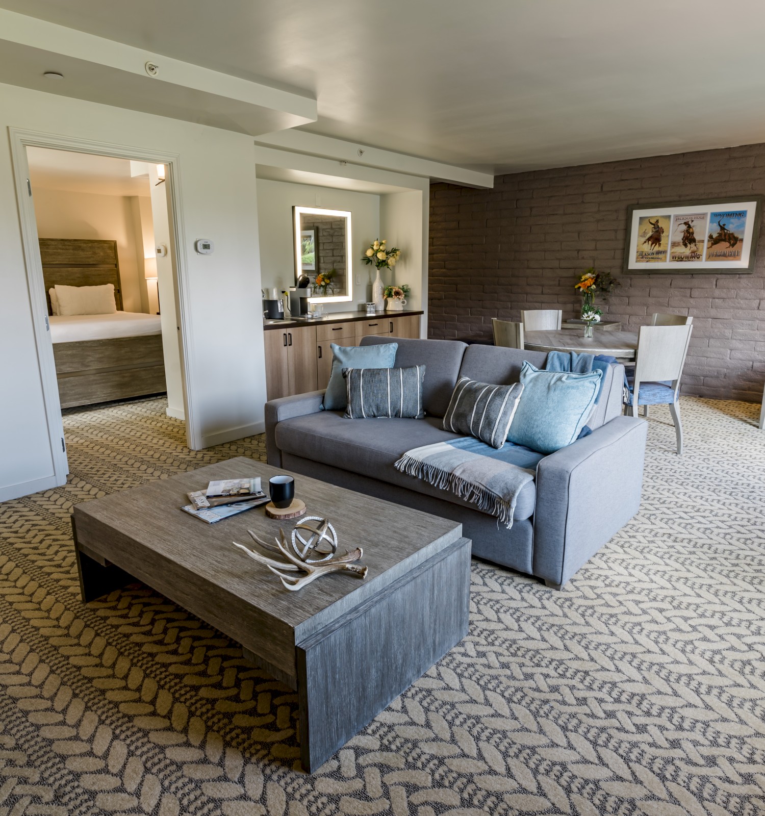 A cozy living room features a gray sofa, coffee table, dining area, and art on brown brick walls. Adjacent is a bedroom with a plush bed.