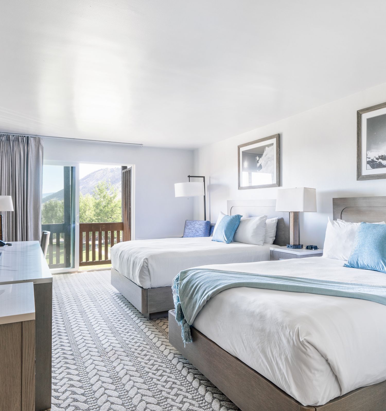 A modern hotel room features two double beds with blue accents, a flat-screen TV on a desk, wall art, and a balcony with a mountain view.