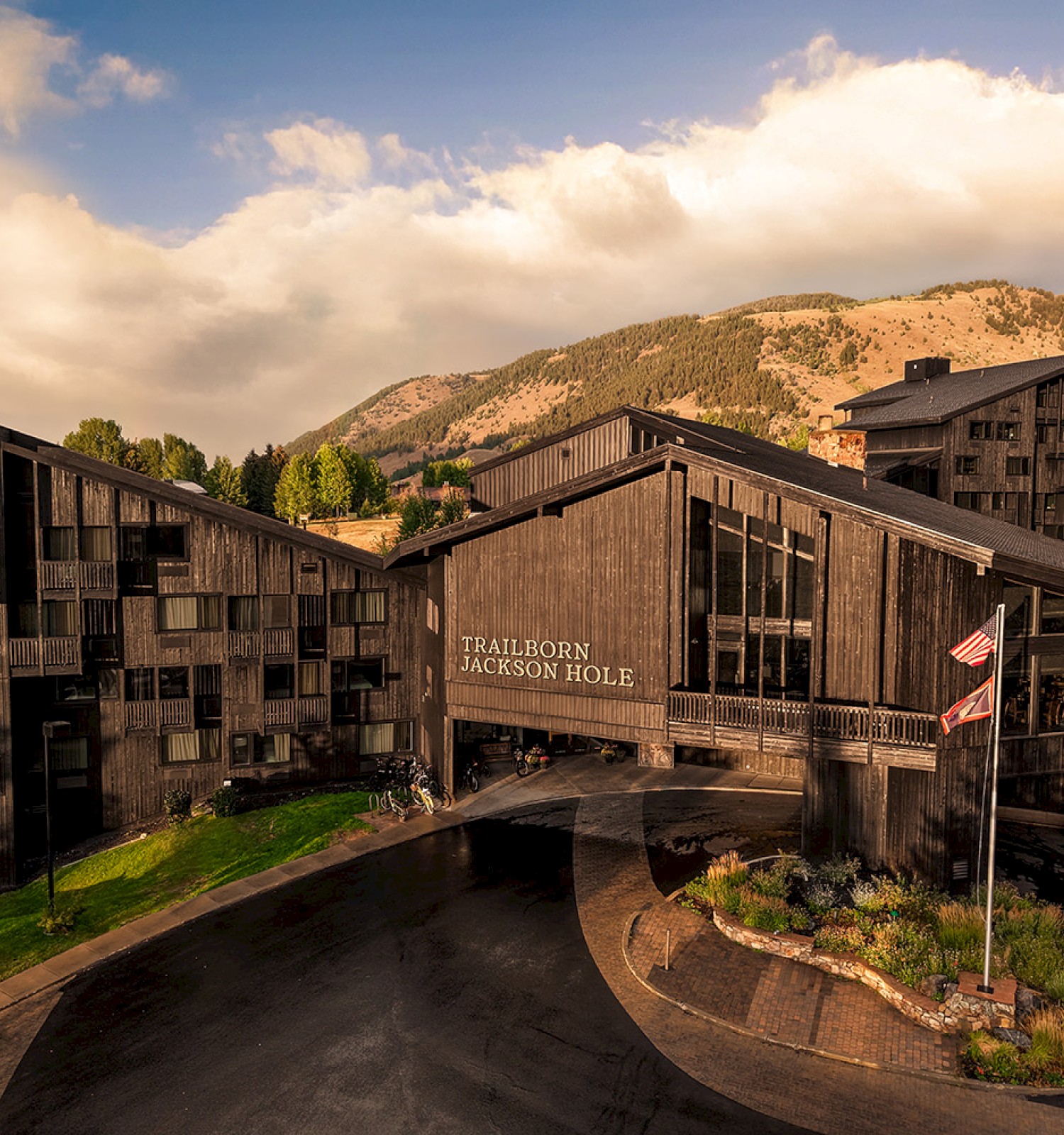 A rustic lodge complex with dark wood buildings, curved driveway, a flag, and a mountainous backdrop under a partly cloudy sky. End.
